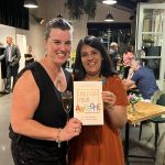 A photo of two women smiling at the camera, holding a copy of the book 'Unleash Your Awesome'. There are other people standing and sitting in the background.