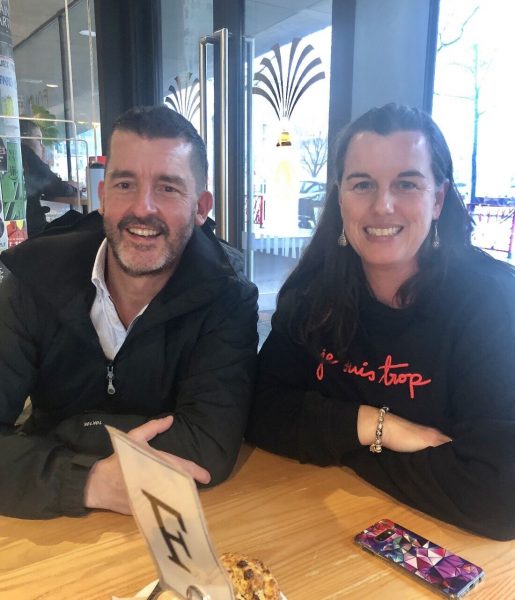 Photo of a brunette man in a dark jacket and white shirt, sitting next to a brunette woman wearing a dark sweatshirt with pink writing. They are smiling at the camera