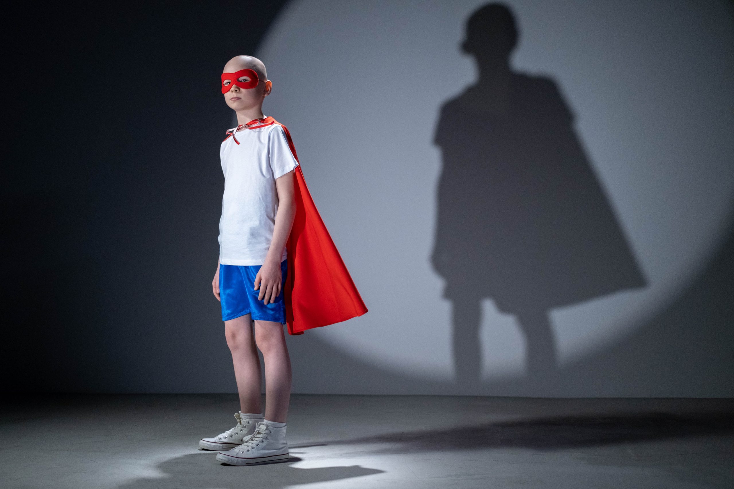 A young child wearing a super-hero cape and amsk, standing in front of a wall with a light projecting a shadow behind them