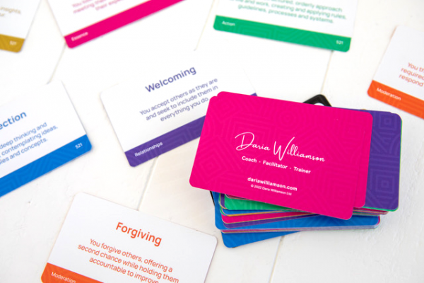 A selection of The Strengths Deck cards laid out on a table with their front, text-based sides facing up, next to a stack of cards that are face down, showing the brightly-coloured reverse sides.