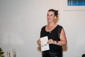 Daria Williamson, a dark-haired woman in a black dress, giving a speech and holding a copy of Unleash Your Awesome