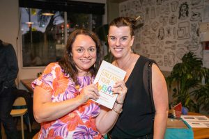 Two women smiling at the camera. One is holding a copy of Unleash Your Awesome.