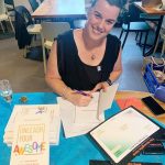Daria Williamson, a dark haired woman in a black dress, sitting at a table and smiling at the camera as she signs a copy of her book, 'Unleash Your Awesome'