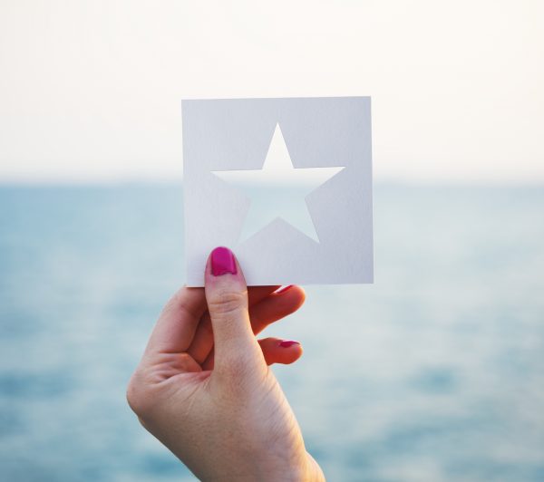 A hand holding a white card in the foreground. The card has a five-pointed star cut out of the middle. In the background is the ocean and horizon line.