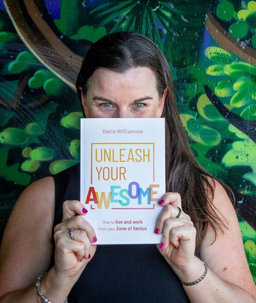 A photo of a dark-haired woman standing in front of a wall painted to resemble a jungle. She is holding a copy of 'Unleash Your Awesome: How to live and work from your Zone of Genius' up so that it is covering the lower half of her face. Her eyes are crinkled in a way that suggests a big smile is hidden behind the book.
