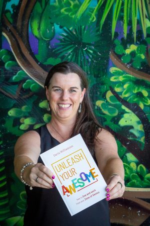 Daria Williamson, a dark-haired woman, is standing in front of a wall painted like a jungle, smiling at the camera. She is holding out a copy of her book "Unleash Your Awesome"
