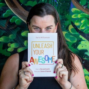 A photo of a dark-haired woman standing in front of a wall painted to resemble a jungle. She is holding a copy of 'Unleash Your Awesome: How to live and work from your Zone of Genius' up so that it is covering the lower half of her face. Her eyes are crinkled in a way that suggests a big smile is hidden behind the book.