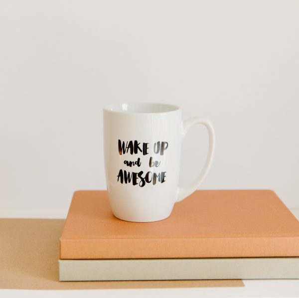 A white coffee mug with the words "Wake up and be awesome" in black. It is on top of two leather-bound books, which are sitting on a wooden table.
