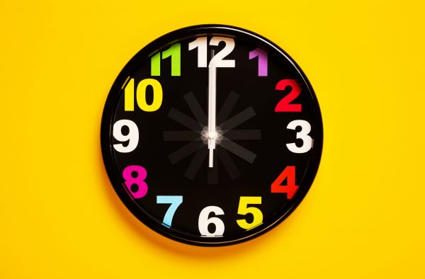 Analogue clock with a black face and different coloured numbers on a yellow wall. One hand is pointing to 12, the other hand is a blur around the clock face