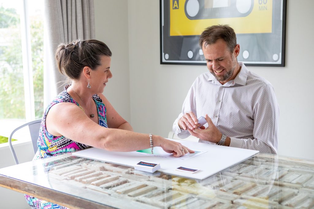 A dark haired woman in a colourful sleeveless dress seated at a table next to a dark haired man in a pale business shirt, holding a set of strengths cards. The woman is looking at the man and pointing at some paper in front of the man, who is looking down at the paper and smiling.
