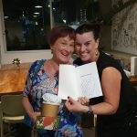 Two women smiling at the camera. One of them is holding up a book, which has a wrong name on the inscription