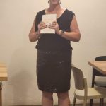 Daria Williamson, a dark haired woman in a black dress, giving a speech and holding a copy of her book, 'Unleash Your Awesome'