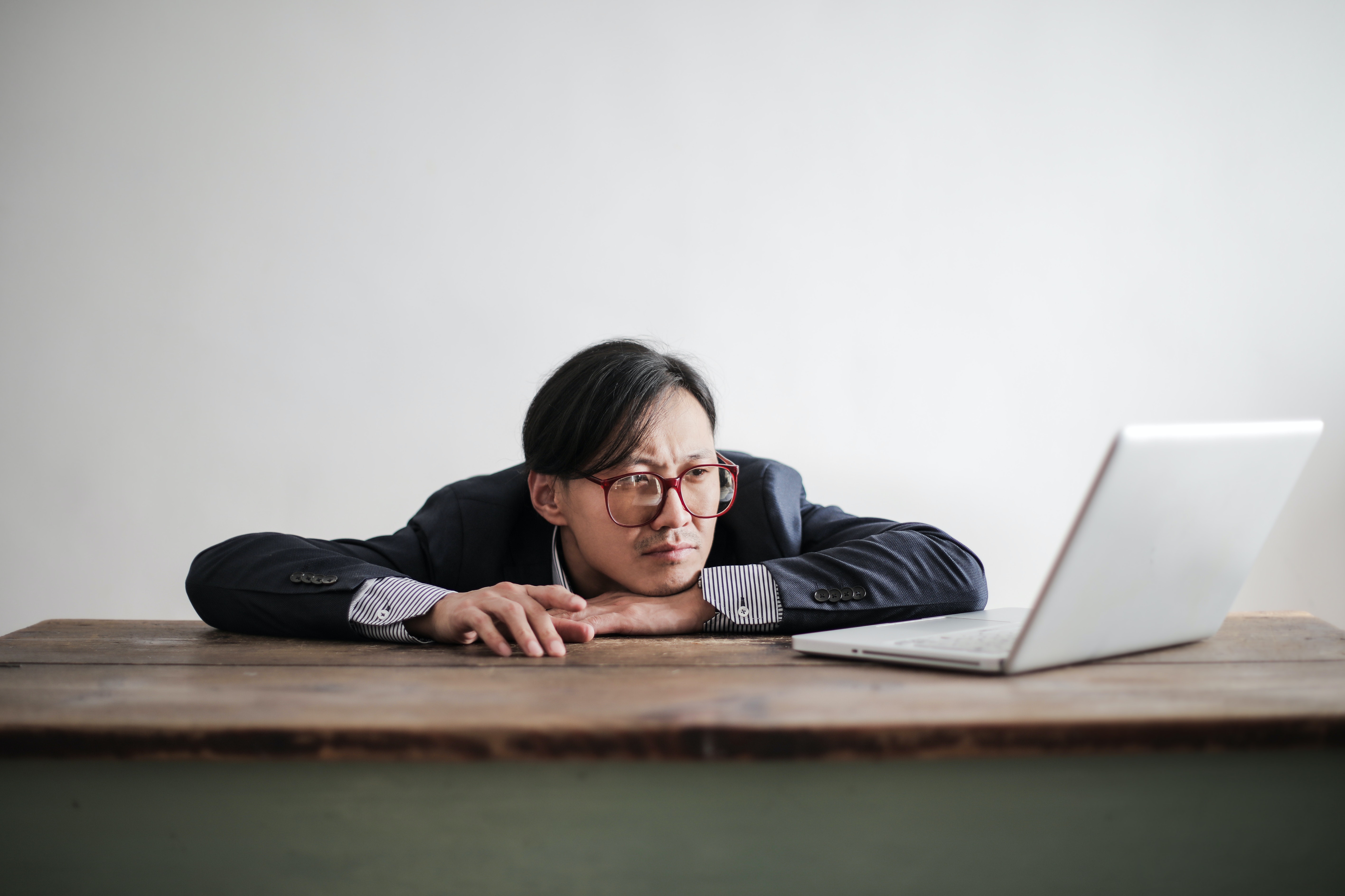 A man wearing glasses and a business suit is slumped on a desk, resting his chin on a hand, with a bored expression on his face. He is looking at an open laptop. He is pointing towards the laptop with one finger of his free hand.