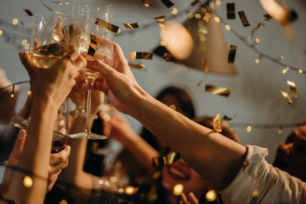 A photo of a group of arms raised, holding champagne glasses, with golden streamers and confetti in the background