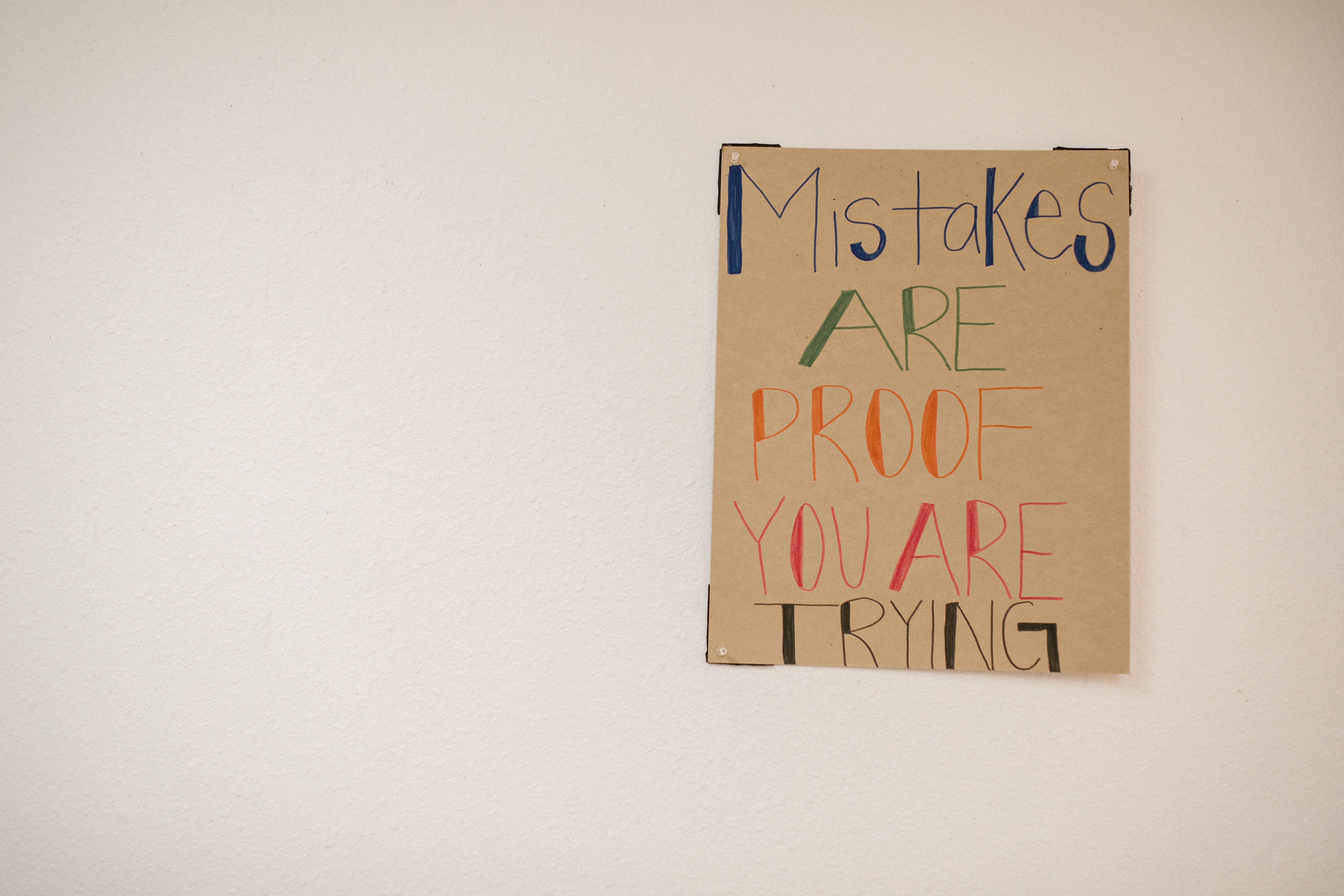 A hand-lettered cardboard sign taped to a wall. The text reads "mistakes are proof you are trying"